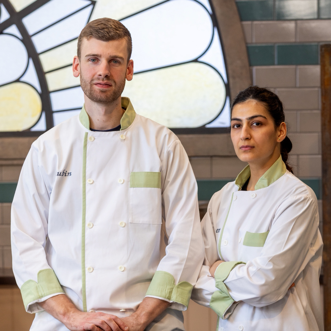 Two chefs in white and light green uniforms stand side by side indoors at Michael Caines at The Stafford, looking at the camera. One chef stands with arms crossed; the other has hands resting in front. A large, decorative window is in the background.