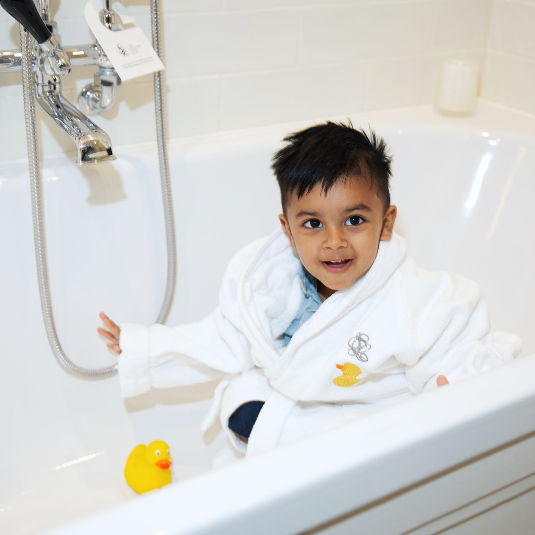 A young child in a white bathrobe sits smiling in a white bathtub next to a yellow rubber duck. The bathroom, which reflects the comfort and elegance found in our room offers, features light-colored tiles and a shiny metal faucet.