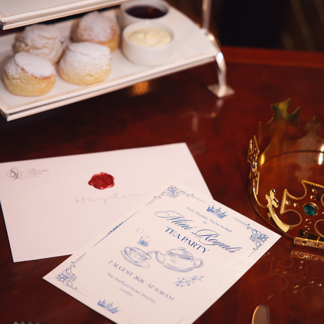 A tea party invitation, a sealed envelope, a gold crown, and a tray with scones and jam are arranged on a polished wooden table, perfectly capturing the charm of afternoon tea.