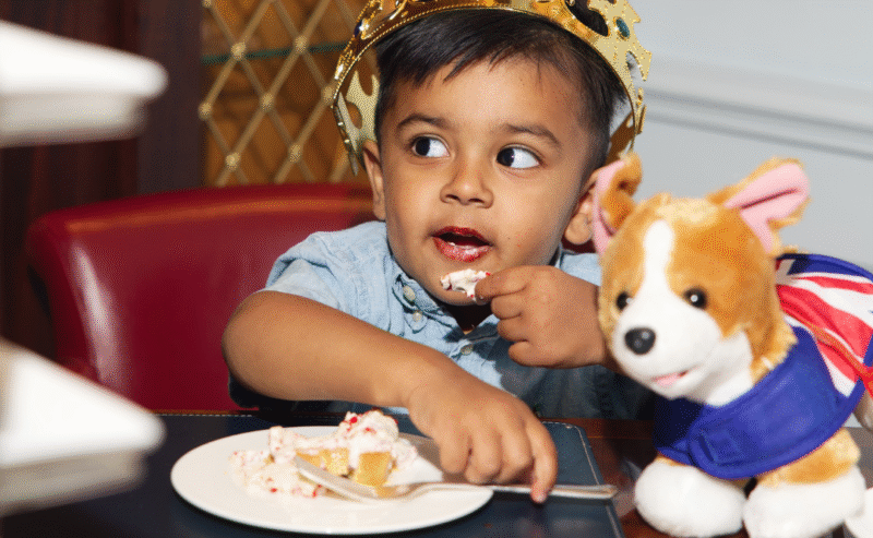 A young child wearing a gold crown enjoys cake at a table during afternoon tea, with a plush corgi toy dressed in a Union Jack cape beside him.