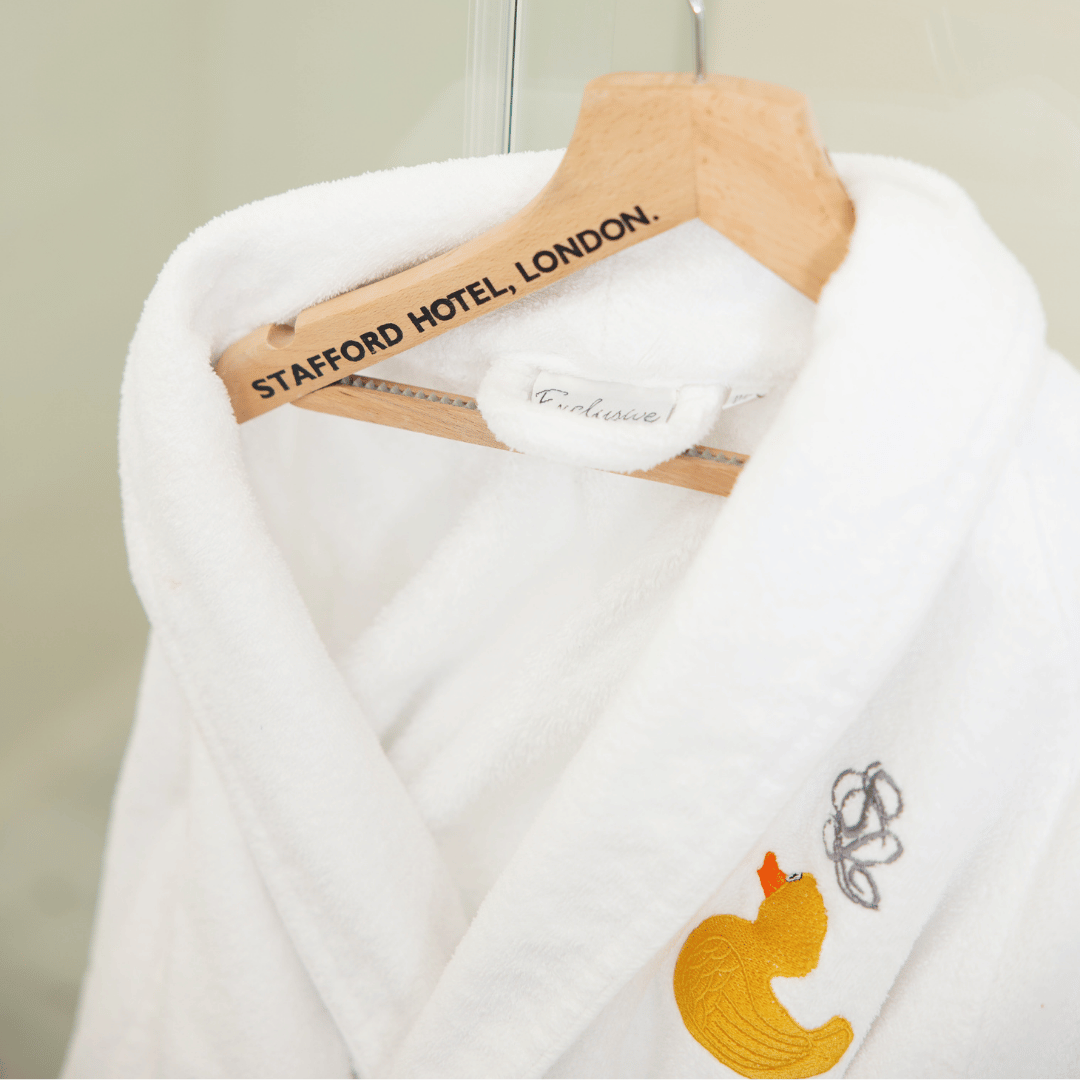 A white bathrobe with gold embroidery hangs on a wooden hanger labeled STAFFORD HOTEL, LONDON, creating an elegant gift idea for Mother’s Day. The background is softly blurred.