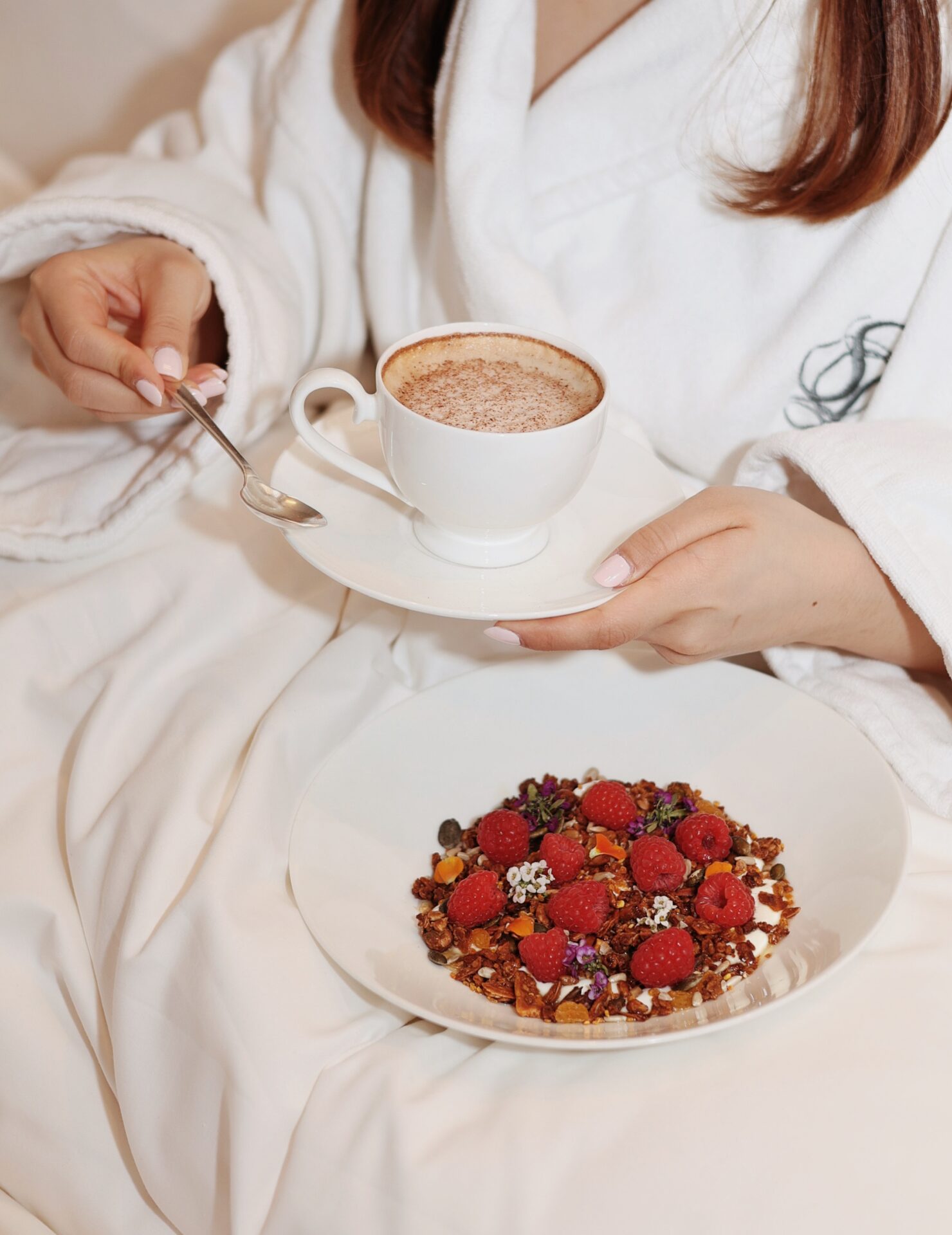 A person in a white robe enjoys In-Room Dining, holding a cup of cappuccino with a saucer and spoon, while a plate of granola topped with raspberries and seeds rests on their lap.