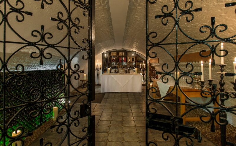 Ornate wrought iron gates open to a wine cellar with stone floors, shelves of wine bottles, a white-clothed table set for wine tasting, flickering candles, and a cozy, arched ceiling.