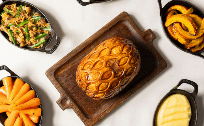A golden-brown pastry on a wooden board is surrounded by small dishes of carrots, mashed potatoes, green beans, squash, parsnips, and stuffing, all arranged neatly on a white surface with decorative gold leaves.