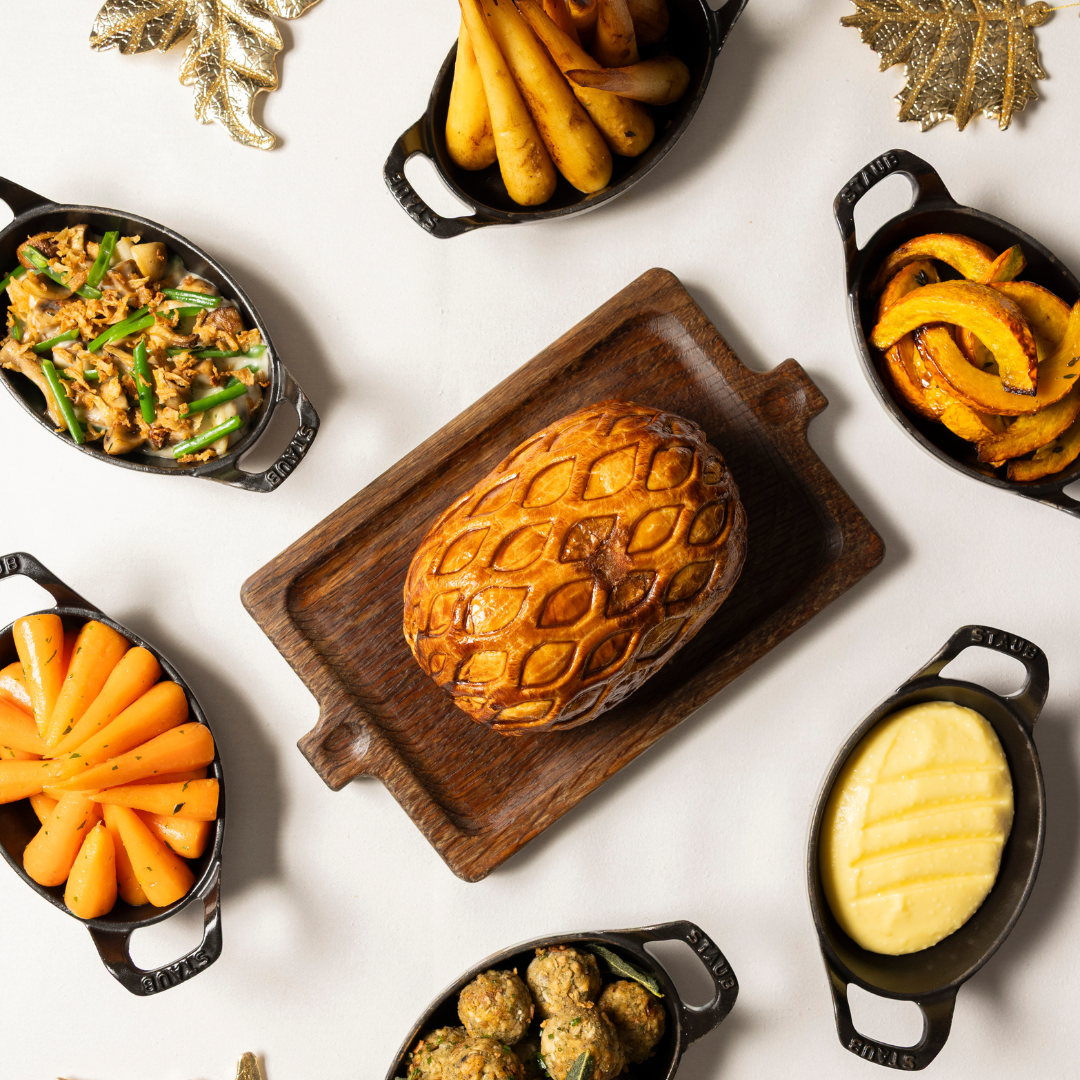 A golden-brown pastry on a wooden board is surrounded by small dishes of carrots, mashed potatoes, green beans, squash, parsnips, and stuffing, all arranged neatly on a white surface with decorative gold leaves.