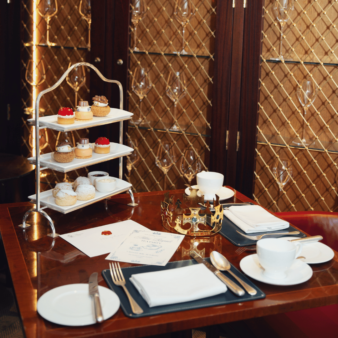 An elegant Mother’s Day afternoon tea setup with pastries on a three-tier stand, two place settings, teacups, utensils, a gold crown, and menus on a polished wooden table with a decorative background.