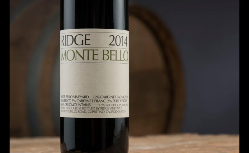 A close-up of a Ridge 2014 Monte Bello wine bottle, showing the label details, set against a blurred background with a wooden barrel—an inviting scene that captures the anticipation of an exceptional wine tasting experience.
