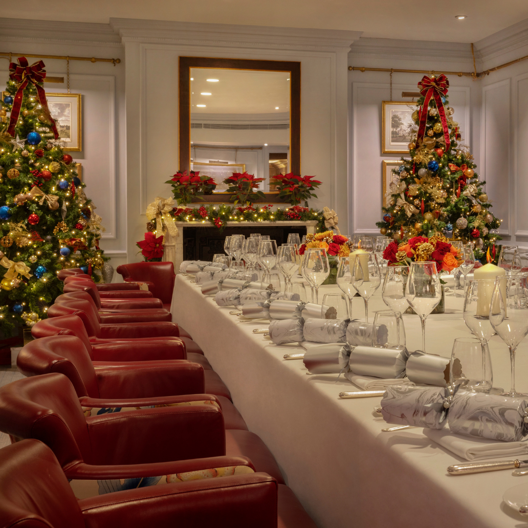 Elegant dining room set for a festive holiday meal, with a long table, red chairs, decorated Christmas trees, wrapped presents, candles, and poinsettias near a fireplace and a large mirror.
