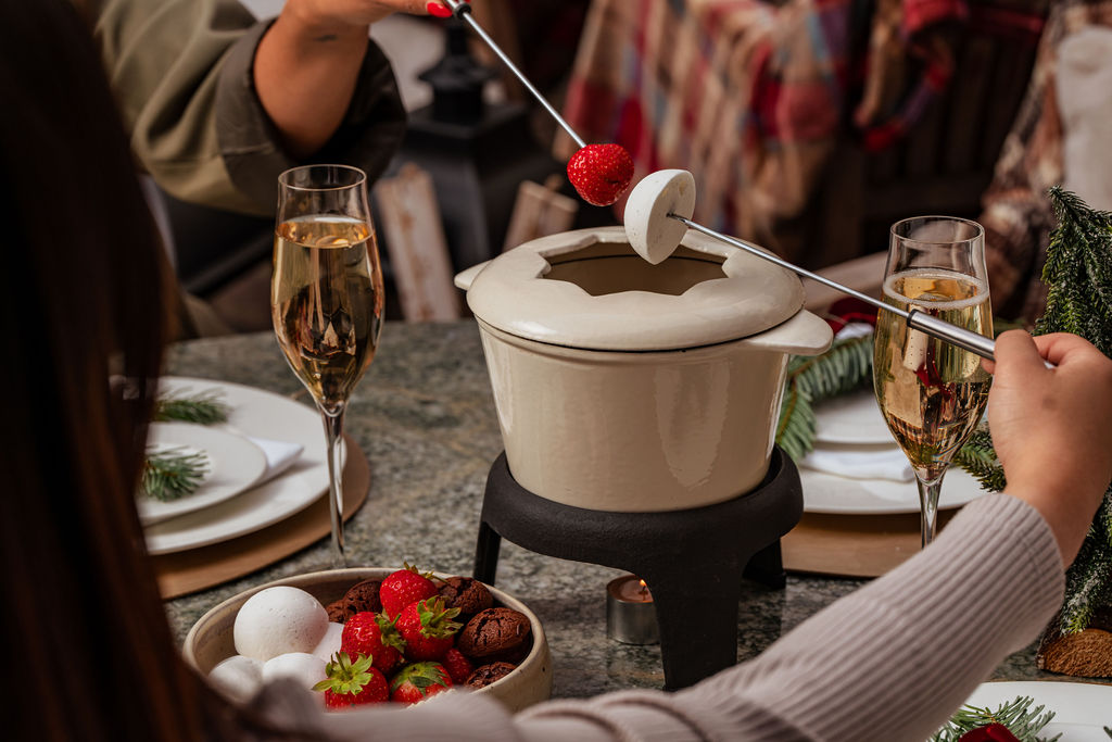 Two people dip a strawberry and a marshmallow into a fondue pot for Mother’s Day, with bowls of fruit and chocolate nearby, next to glasses of champagne.
