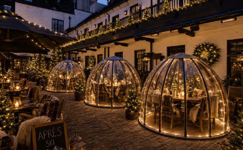 A cosy outdoor courtyard at night, featuring three glass dining domes decorated with fairy lights for Christmas, surrounded by festive greenery and warm glowing lanterns. A chalkboard sign reads Après Ski.