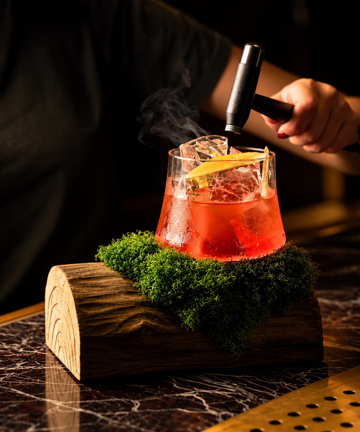 A hand uses a small blowtorch to smoke a Mother’s Day red cocktail, garnished with a lemon twist and served on a moss-covered log.