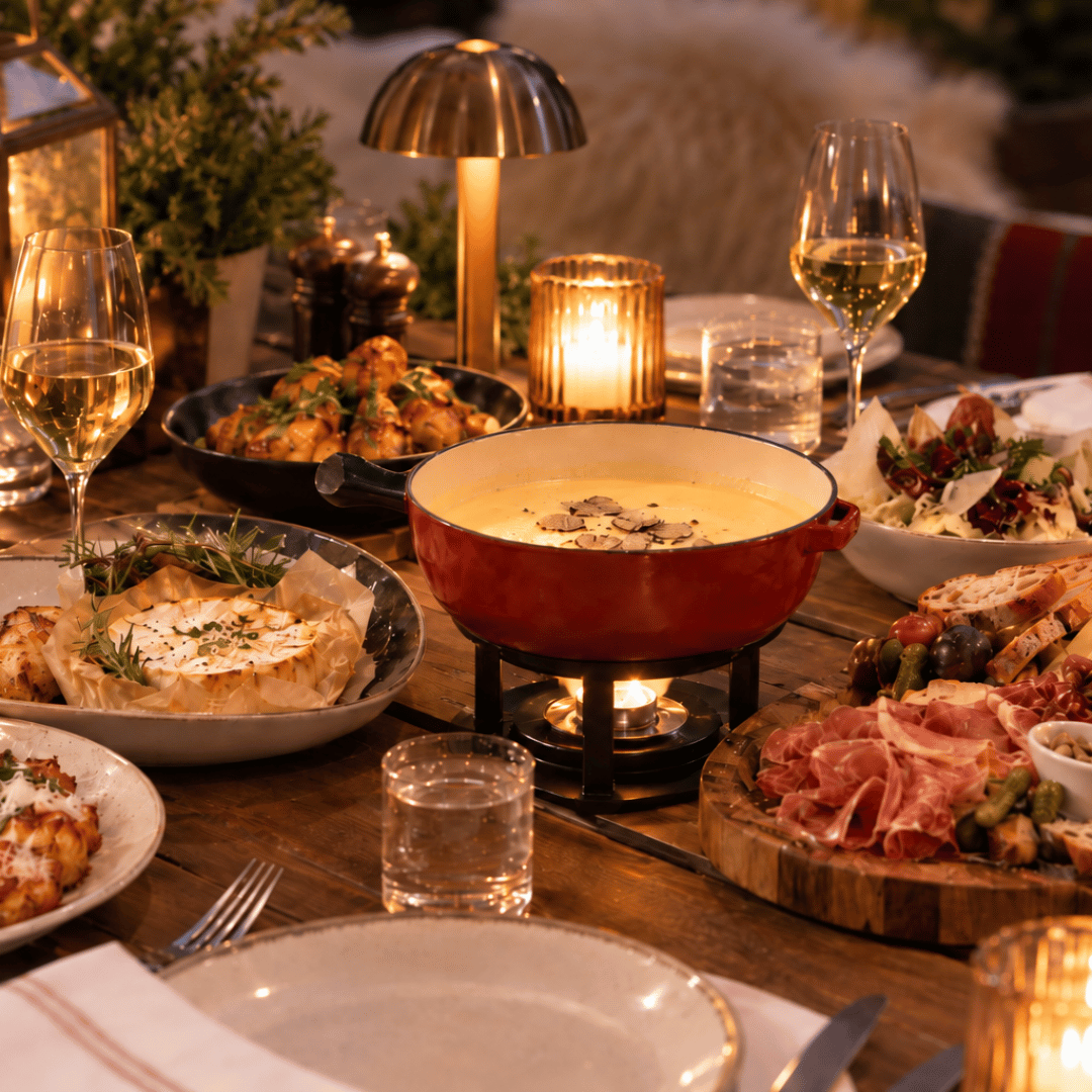 A cosy Mother’s Day dinner table set with cheese fondue, charcuterie, salad, roasted vegetables, bread, and white wine, surrounded by lit candles and warm décor.
